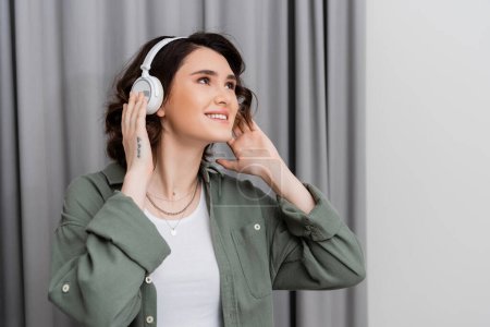 Photo for Delighted young woman in casual clothes, with tattoo and with wavy brunette hair looking away while listening music in wireless headphones near grey curtains in modern hotel room, weekend getaway - Royalty Free Image