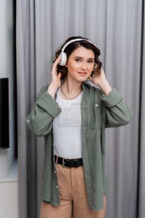 pleased woman with wavy brunette hair and tattoo looking at camera while listening podcast in wireless headphones near grey curtains in modern hotel suite, leisure, weekend getaway