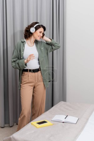 mujer tatuada sin preocupaciones con cabello moreno ondulado escuchando música en auriculares inalámbricos mientras está de pie cerca de cortinas grises, teléfono inteligente con pantalla en blanco y cuadernos en la cama en una cómoda suite de hotel