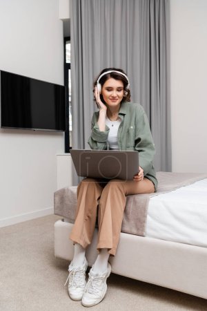 glückliche junge Frau mit welligem brünetten Haar und Tätowierung, die mit Laptop in der Nähe des LCD-Fernsehers auf dem Bett sitzt, grauen Vorhängen, Einstellen drahtloser Kopfhörer während des Webinars in der Hotelsuite, im Studium und auf Reisen