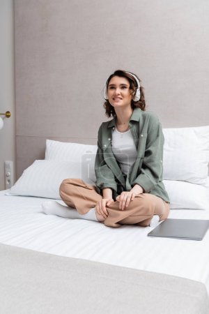 joyful woman with brunette hair, in casual clothes and headphones sitting on bed with crossed legs and listening musical podcast in wireless headphones near white pillows and laptop in hotel room