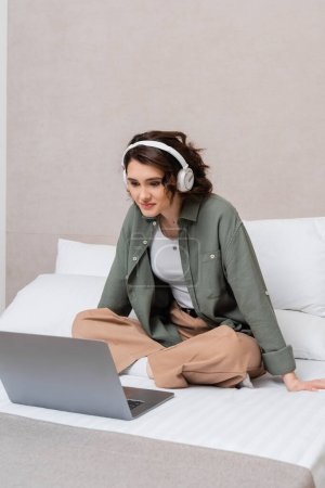 femme souriante avec cheveux bruns ondulés, en vêtements décontractés et écouteurs sans fil assis sur le lit près des oreillers blancs et regarder des films sur ordinateur portable dans la suite moderne de l'hôtel, les loisirs et les voyages