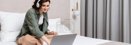 pleased young woman with wavy brunette hair, in wireless headphones watching movie on laptop while sitting on bed with crossed legs near grey curtains and wall sconce in hotel room, banner