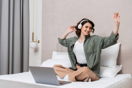 überglückliche tätowierte Frau mit welligem brünetten Haar und geschlossenen Augen, die mit überkreuzten Beinen neben Laptop, Kissen und Wandleuchte auf dem Bett sitzt, Musik-Podcast hört und im modernen Hotelzimmer gestikuliert