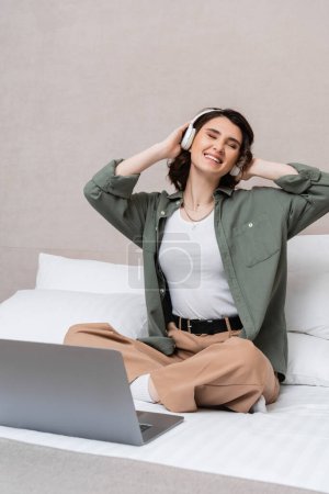 carefree woman with wavy brunette hair and closed eyes listening musical podcast in wireless headphones while sitting on bed near laptop and white pillows in hotel suite, leisure and travel