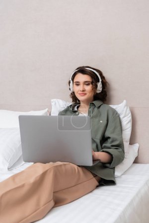 jeune femme aux cheveux bruns ondulés, en vêtements décontractés et écouteurs sans fil assis sur le lit près d'oreillers blancs et souriant tout en regardant un film sur ordinateur portable dans la chambre d'hôtel 