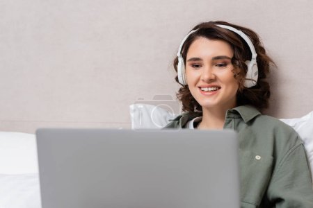 young and cheerful woman with wavy brunette hair watching film on blurred laptop while sitting in wireless headphones near grey wall and white pillows in modern hotel room
