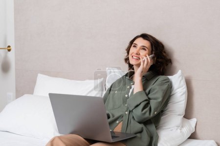 Photo for Smiling woman with wavy brunette hair wearing casual clothes, holding laptop and talking on mobile phone while sitting on bed near white pillows in cozy hotel room, work-life integration - Royalty Free Image