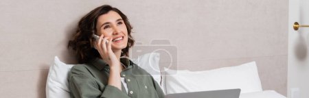 mujer llena de alegría con el pelo ondulado morena y tatuaje hablando en el teléfono móvil cerca de la computadora portátil, almohadas blancas y la pared gris en la suite de hotel moderna, estilo de vida independiente, trabajo y viajes, pancarta