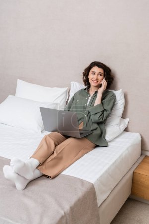 full length of young and happy woman in casual clothes, with wavy brunette hair looking away while sitting on bed with laptop and talking on mobile phone near white pillows in hotel suite