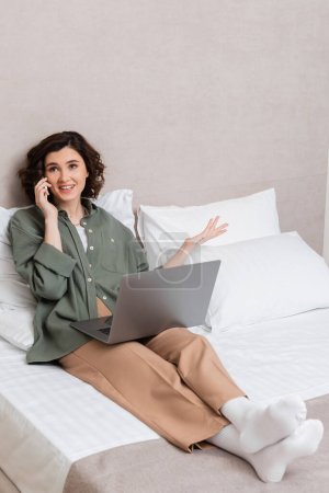 fröhliche junge Frau mit welligem brünetten Haar und Tätowierung, in lässiger Kleidung gestikulierend während eines Gesprächs auf dem Smartphone, während sie mit Laptop neben weißen Kissen im Hotelzimmer sitzt