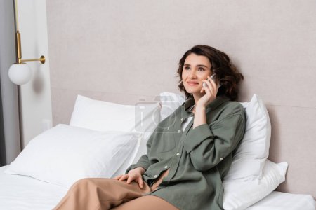 pleased young woman in casual clothes, with wavy brunette hair talking on mobile phone near grey wall and white pillows on comfortable bed in model hotel room, leisure and travel