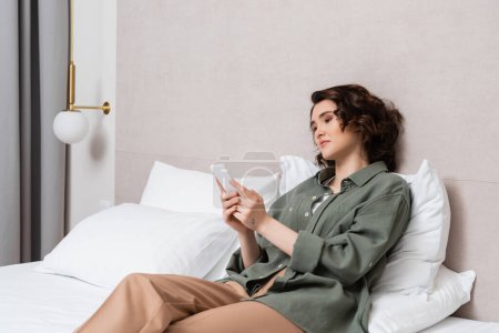 young woman with wavy brunette hair and tattoo sitting on bed in casual closed and browsing internet on mobile phone near white pillows and wall sconce in comfortable hotel room, leisure and travel