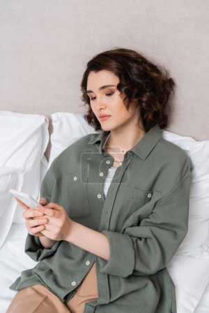 young woman with wavy brunette hair and tattoo browsing internet on mobile phone while sitting on bed near grey wall and white pillows in cozy atmosphere of hotel room, leisure and travel