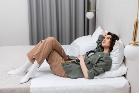 Photo for Full length of dreamy and carefree woman with wavy brunette hair laying on bed in casual clothes near white pillows, wall sconces and grey curtains in cozy atmosphere of modern hotel room - Royalty Free Image