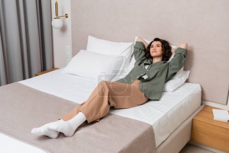 full length of young delighted woman with wavy brunette hair lying on bed and white pillows in casual clothes near bedside table, books and wall sconce in contemporary hotel room