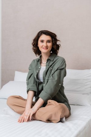 carefree woman with wavy brunette hair sitting on bed in casual clothes, with crossed legs, looking at camera near white pillows and grey wall in contemporary hotel suite, leisure and travel