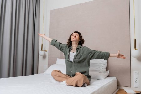 young excited woman with wavy brunette hair and in casual clothes sitting on bed with outstretched hands and looking up near white pillows, wall sconces and grey curtains in comfortable hotel room