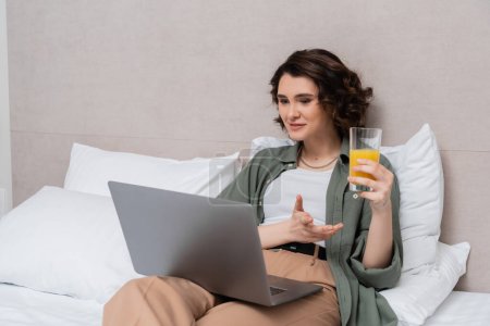 lächelnde Frau mit welligem brünetten Haar hält ein Glas frischen Orangensaft in der Hand und gestikuliert beim Videoanruf auf dem Laptop, während sie auf dem Bett neben weißen Kissen und grauen Wänden im Hotelzimmer sitzt