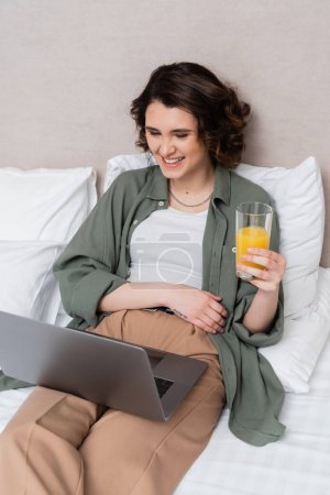 mujer joven y despreocupada en ropa casual, con el pelo ondulado morena y tatuaje sosteniendo vaso de jugo de naranja fresca mientras ve la película en el ordenador portátil en la cama cerca de almohadas blancas y la pared gris en la habitación de hotel