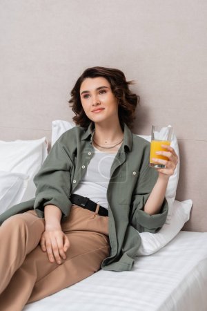 femme ravie et rêveuse aux cheveux bruns ondulés tenant un verre de jus d'orange frais tout en étant assise sur un lit près d'oreillers blancs et d'un mur gris dans une suite hôtelière moderne, loisirs et voyages