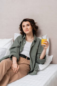 delighted and dreamy woman with wavy brunette hair holding glass of fresh orange juice while sitting on bed near white pillows and grey wall in modern hotel suite, leisure and travel Poster #659229700