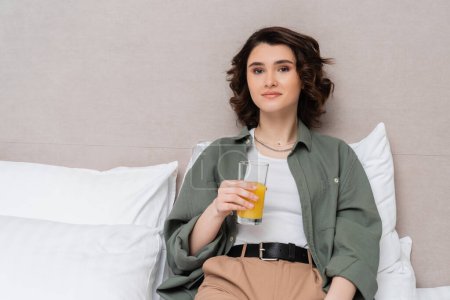 pleased woman in casual clothes, with wavy brunette hair sitting on bed with glass of fresh orange juice and looking at camera near white pillows and grey wall in cozy hotel room