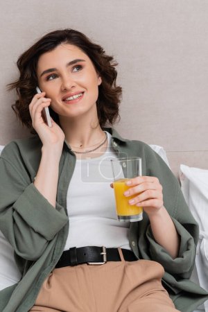 happy woman in casual clothes, with wavy brunette hair talking on mobile phone on bed near white pillows and grey wall in cozy atmosphere of hotel suite, leisure and travel