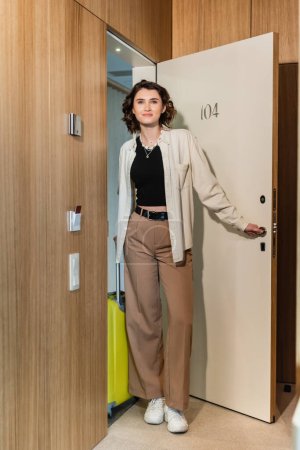 full length of pleased woman in white shirt and beige pants, with wavy brunette hair holding yellow travel bag and smiling while opening door of hotel suite near keycard reader, check-in 