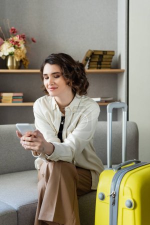 entzückte tätowierte Frau mit welligem brünetten Haar sitzt auf der Couch neben der gelben Reisetasche und surft im Internet auf dem Smartphone in der Nähe von Büchern und Vase mit Blumen auf verschwommenem Hintergrund in der Hotellobby