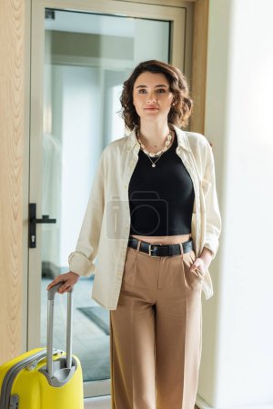 Photo for Young woman in stylish casual clothes, white shirt, black crop top and beige pants standing with hand in pocket near yellow travel bag and glass door of contemporary hotel, travel lifestyle - Royalty Free Image