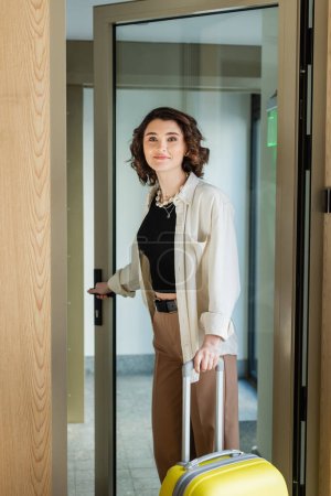 delighted woman with wavy brunette hair, in white shirt, black crop top and beige pants holding yellow suitcase, opening glass door and entering lobby of modern hotel, travel lifestyle