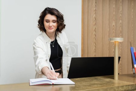 friendly concierge in stylish casual clothes, with wavy brunette hair writing in notebook near computer monitor and lamp while working at reception desk in lobby of contemporary hotel