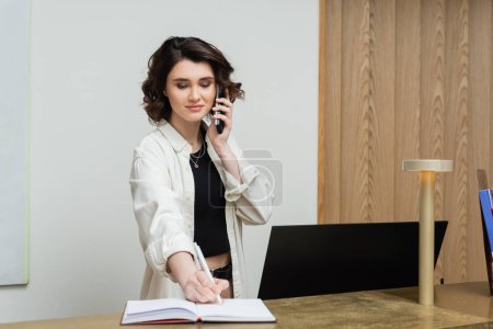 Photo for Joyful receptionist with wavy brunette hair working at front desk, writing in notebook and talking on telephone near computer monitor in lobby of modern hotel, hospitality industry - Royalty Free Image