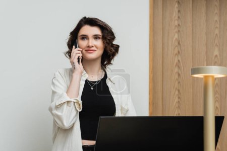 pleased young woman in stylish casual clothes, with wavy brunette hair and tattoo talking on telephone while working as receptionist at front desk near computer monitor and lamp in hotel lobby