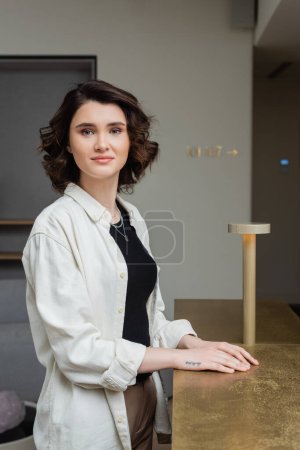young and joyful woman with wavy brunette hair, in trendy casual clothes, white shirt and black crop top looking at camera near reception desk and lamp in lobby of contemporary hotel