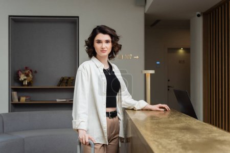 confident woman in black crop top, white shirt and beige pants, with wavy brunette hair standing near lamp on reception desk and looking at camera in lobby of modern hotel, travel lifestyle