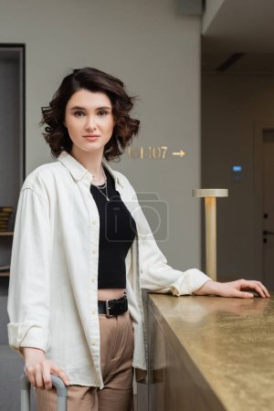 front desk, young and fashionable woman with wavy brunette hair, in white shirt, black crop top and beige pants looking at camera while standing near reception desk and lamp in modern hotel 
