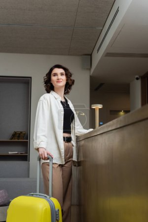 young and trendy woman with short brunette hair, in white shirt, black crop top and beige pants standing with yellow suitcase near reception desk and looking away in lobby of modern hotel