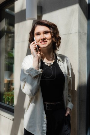 mujer tatuada feliz con el pelo ondulado morena, vistiendo camisa blanca, top negro y pantalones cortos mientras habla en el teléfono móvil en la luz del sol cerca del edificio de hotel moderno en la calle de la ciudad