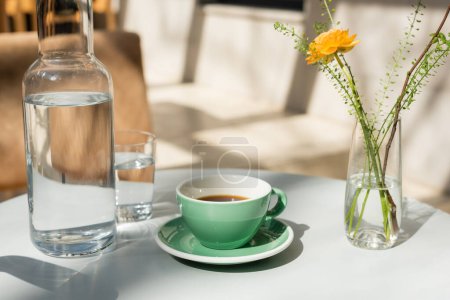 vase with yellow rose and green plants, glass and decanter with fresh pure water, saucer, cup with black coffee on white round table in morning sunlight, outdoor terrace of hotel cafe
