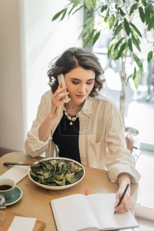 jeune femme tatouée avec des cheveux bruns courts parler sur smartphone et écrire dans un carnet près de salade de légumes et tasse de café aromatique sur la table dans le hall café de l'hôtel moderne, le travail et les voyages