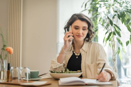 smiling woman with wavy brunette hair and tattoo talking on mobile phone and writing in notebook near vegetable salad and cup of aromatic coffee in lobby cafe of modern hotel, freelance lifestyle