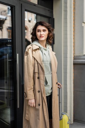 attraktive und stilvolle Frau in beigem Trenchcoat und grauem Kapuzenpullover, die wegschaut, während sie mit gelber Reisetasche am Eingang eines modernen Hotels in der Straße der europäischen Stadt steht