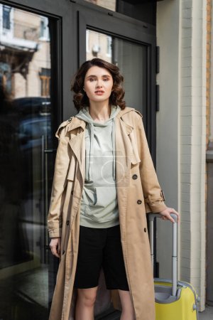 Photo for Confident woman with wavy brunette hair, in grey hoodie and beige trench coat standing with yellow suitcase and looking at camera near glass doors of modern hotel on city street, travel lifestyle - Royalty Free Image