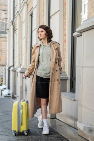 full length of woman with wavy brunette hair, in beige trench coat, grey hoodie, black shorts and white sneakers walking with yellow suitcase along building on urban street of European city