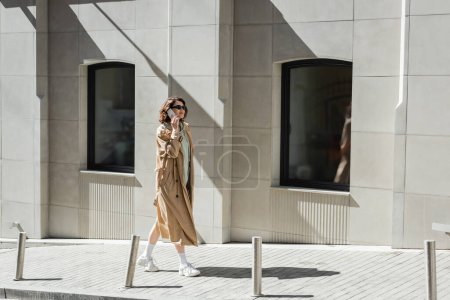 eine junge, modische Frau in dunkler Sonnenbrille, beigem Trenchcoat und weißen Turnschuhen, die mit dem Smartphone spricht, während sie durch ein modernes Gebäude auf der Straße in der europäischen Stadt läuft 