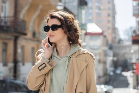 modische tätowierte Frau mit dunkler Sonnenbrille, grauem Kapuzenpulli und beigem Trenchcoat, die bei einem Handy-Gespräch in der Stadt auf verschwommenem Hintergrund wegschaut, Straßenfotografie