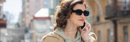 portrait of young tattooed woman with wavy brunette hair wearing dark sunglasses, beige trench coat and grey hoodie talking on mobile phone on blurred street of european city, banner