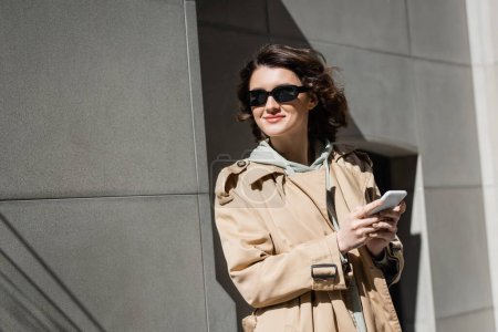 zufriedene und trendige Frau mit dunkler Sonnenbrille, Kapuzenpulli und beigem Trenchcoat, die auf dem Smartphone Nachrichten sendet und wegschaut, während sie in der Nähe eines grauen Gebäudes in der Stadt steht, urbaner Lifestyle, Straßenfotografie